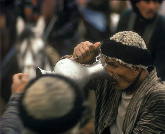 A horseman in a fur-trimmed cap and striped quilted robe drinking from a metal jug in northern Afghanistan, photographed by Herbert Maeder in 1968