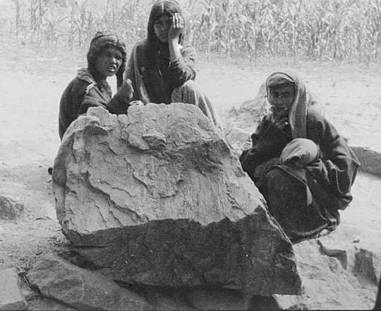 Three Kati women in wrapped shawls resting beside a boulder near Dageri village, Lutkoh valley, Chitral, 1929