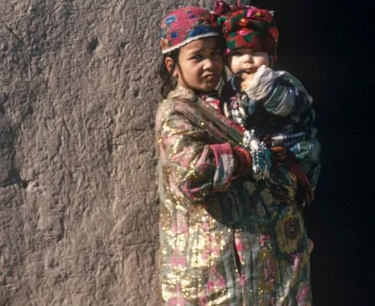 A girl in a patterned robe and embroidered cap holding a baby beside a house in Mazar-i-Sharif, Balkh, photographed by Herbert Maeder in 1968