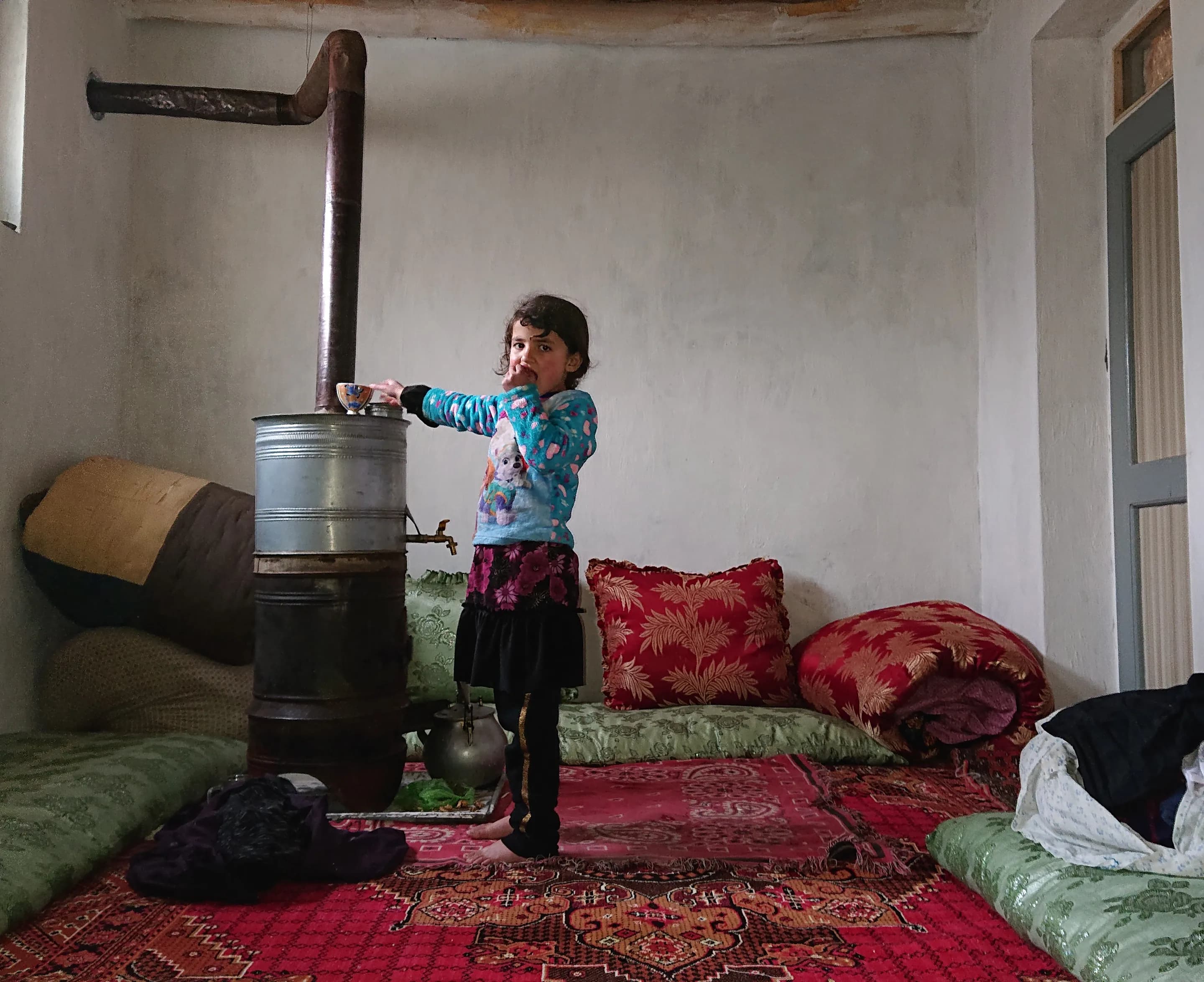 A young girl warming herself beside a handmade stove in a home in Warsaj, Takhar